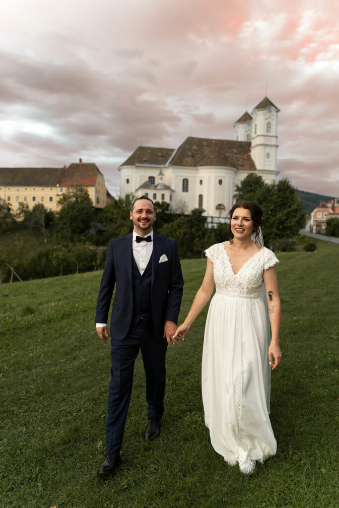 Brautpaar auf der Wiese vor der Weizbergkirche, Hand in Hand.