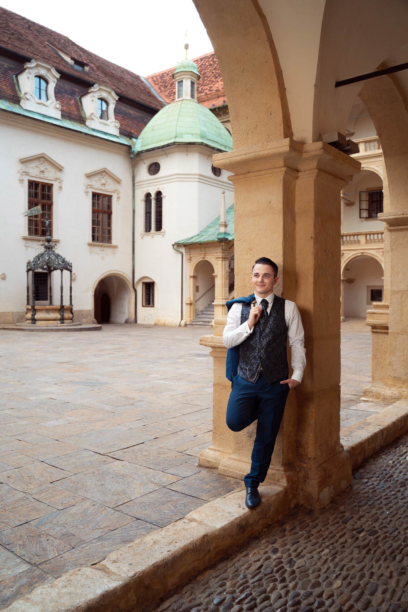 Der Bräutigam lehnt beim After Wedding Shooting lässig an einer Säule, im Hintergrund sieht man ein altes Gebäude mit Kupferdach. Er trägt einen dunkelblauen Anzug mit Veste und hat sich sein Sakko locker über die Schulter gehängt.