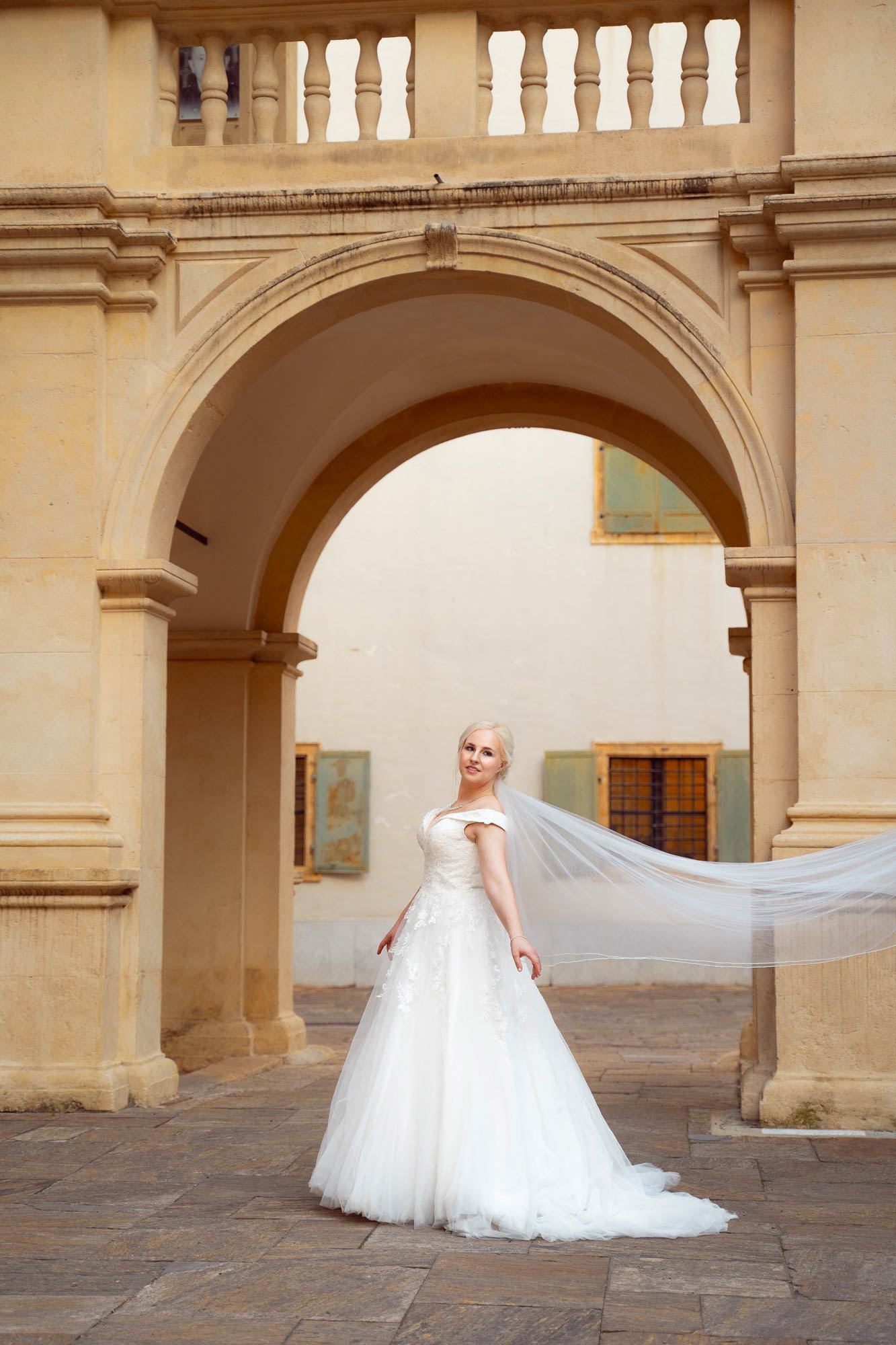 Die Braut steht in einem langen weißen Kleid mit Reifrock vor einem gelben Torbogen, ihr langer, weißer Schleier weht im Wind.