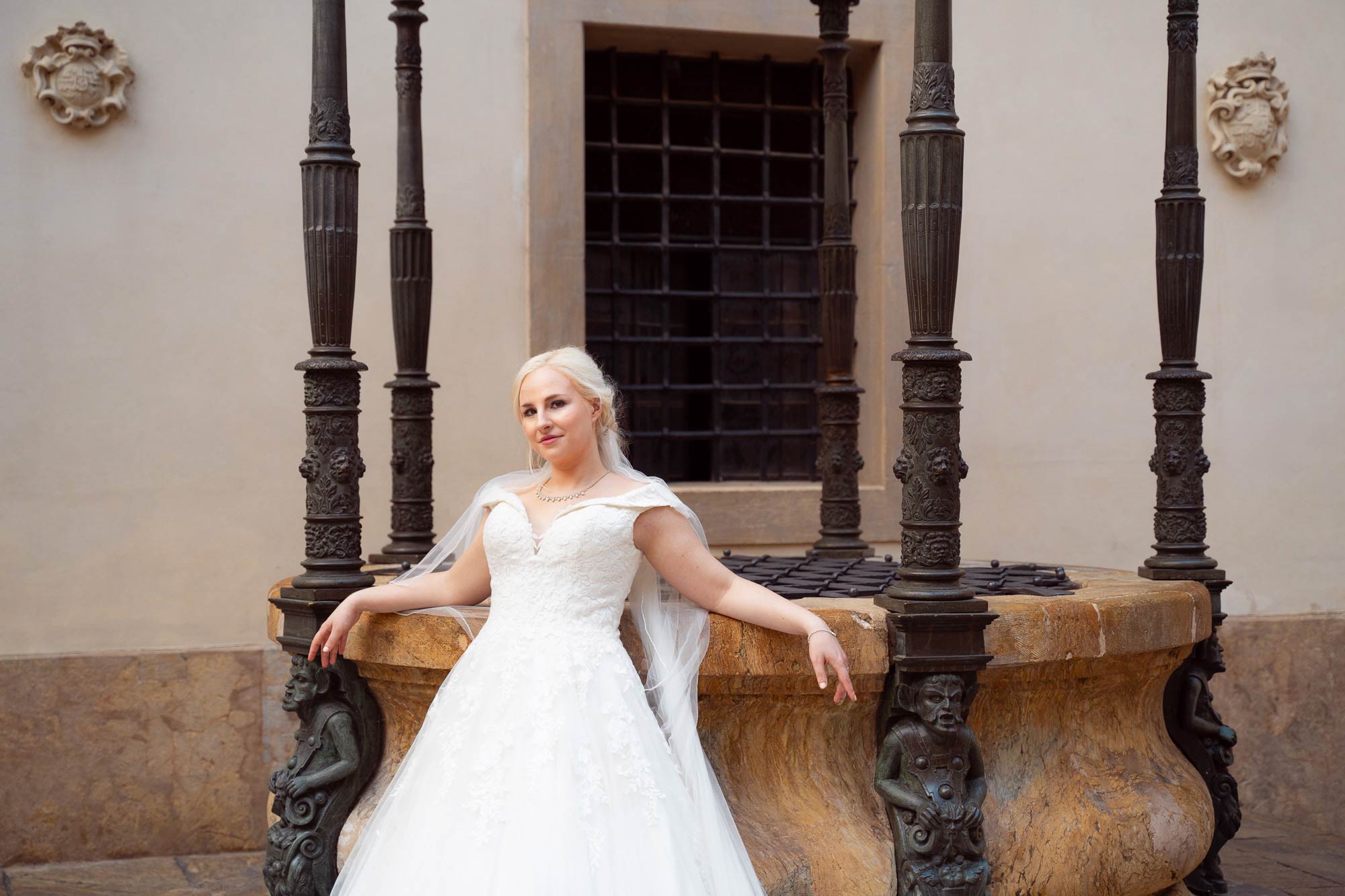 Braut in langem weißen Kleid, lehnt sich an einen mittelalterlichen Brunnen und schaut verträumt in die Luft.
