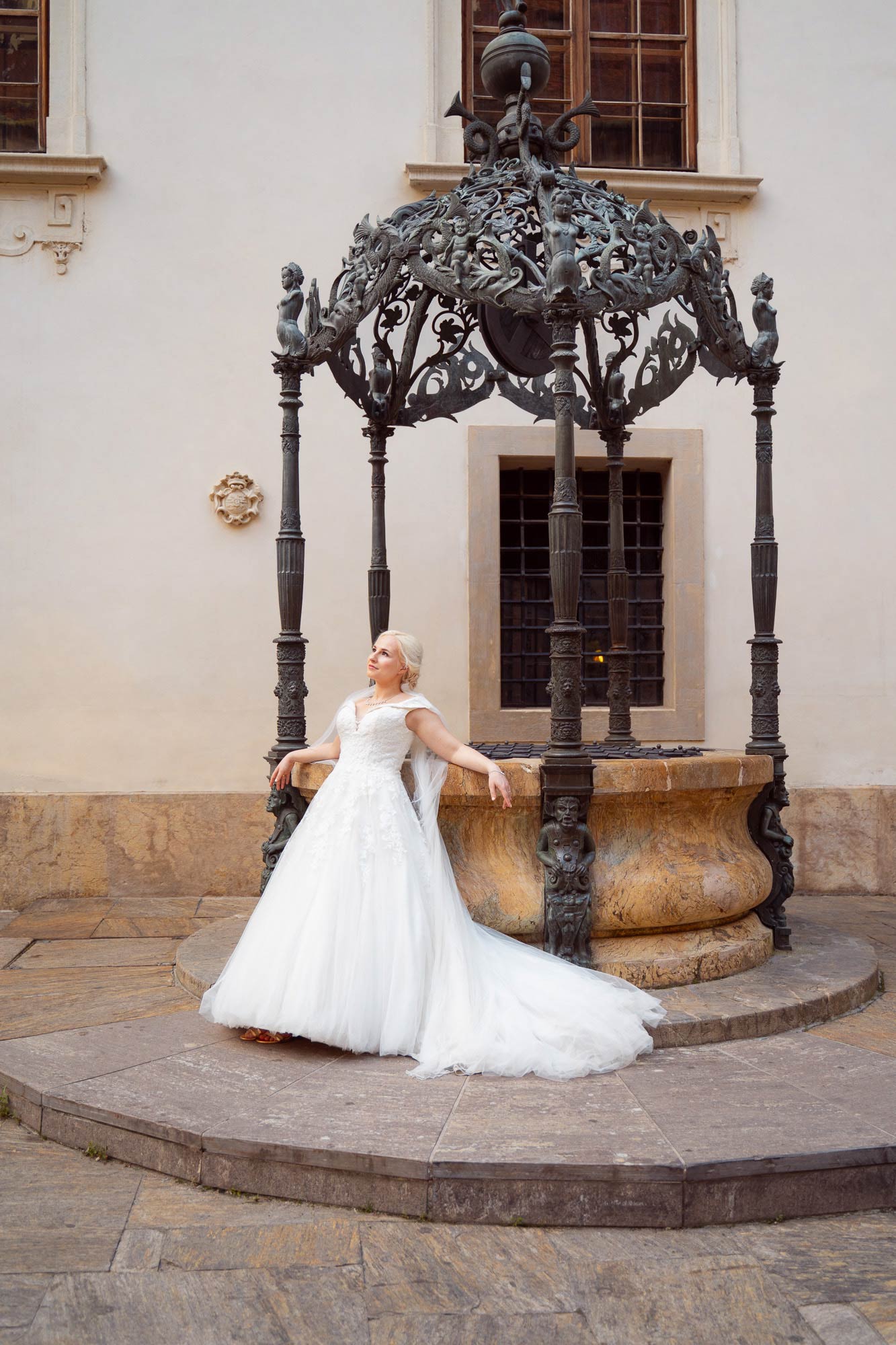 Braut in langem weißen Kleid, lehnt sich an einen mittelalterlichen Brunnen und schaut verträumt in die Luft.